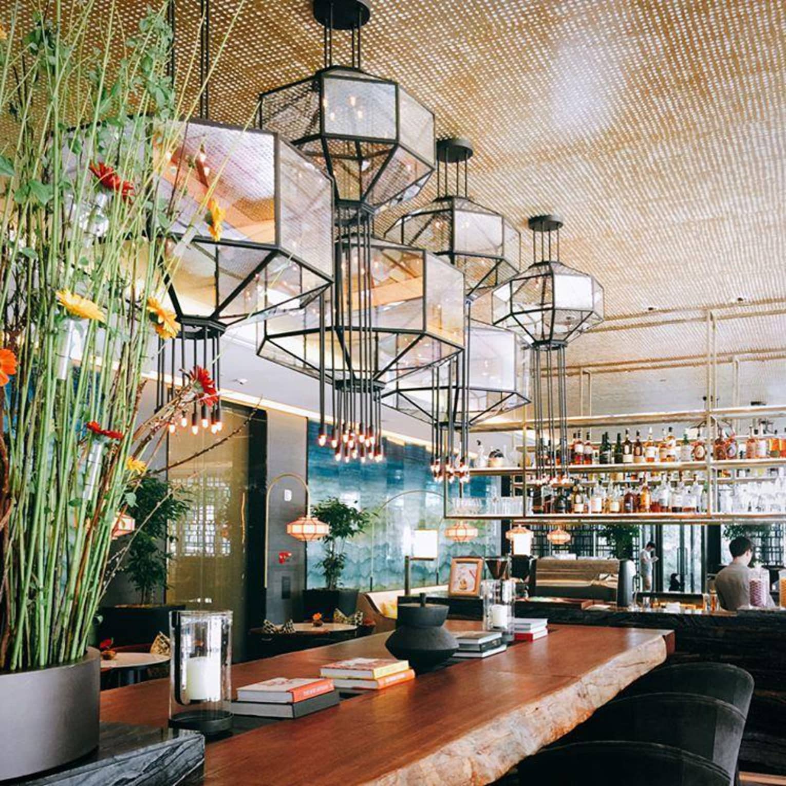 Modern glass lanterns hang over rustic wood bar with books, flowers, vases in sunny lounge